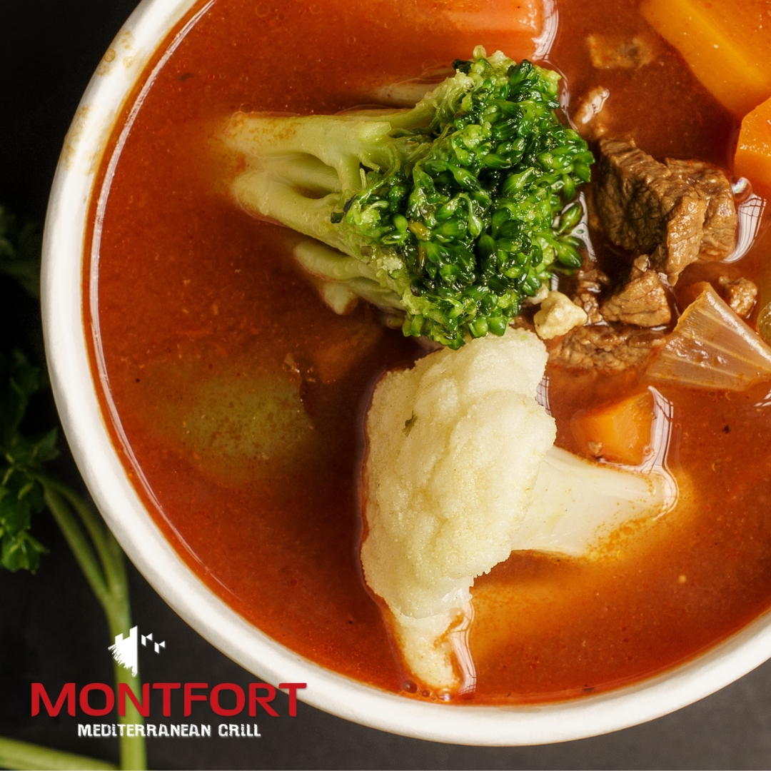 Bowl of vegetable and beef soup with broccoli, cauliflower, carrots, and onions in a reddish broth.