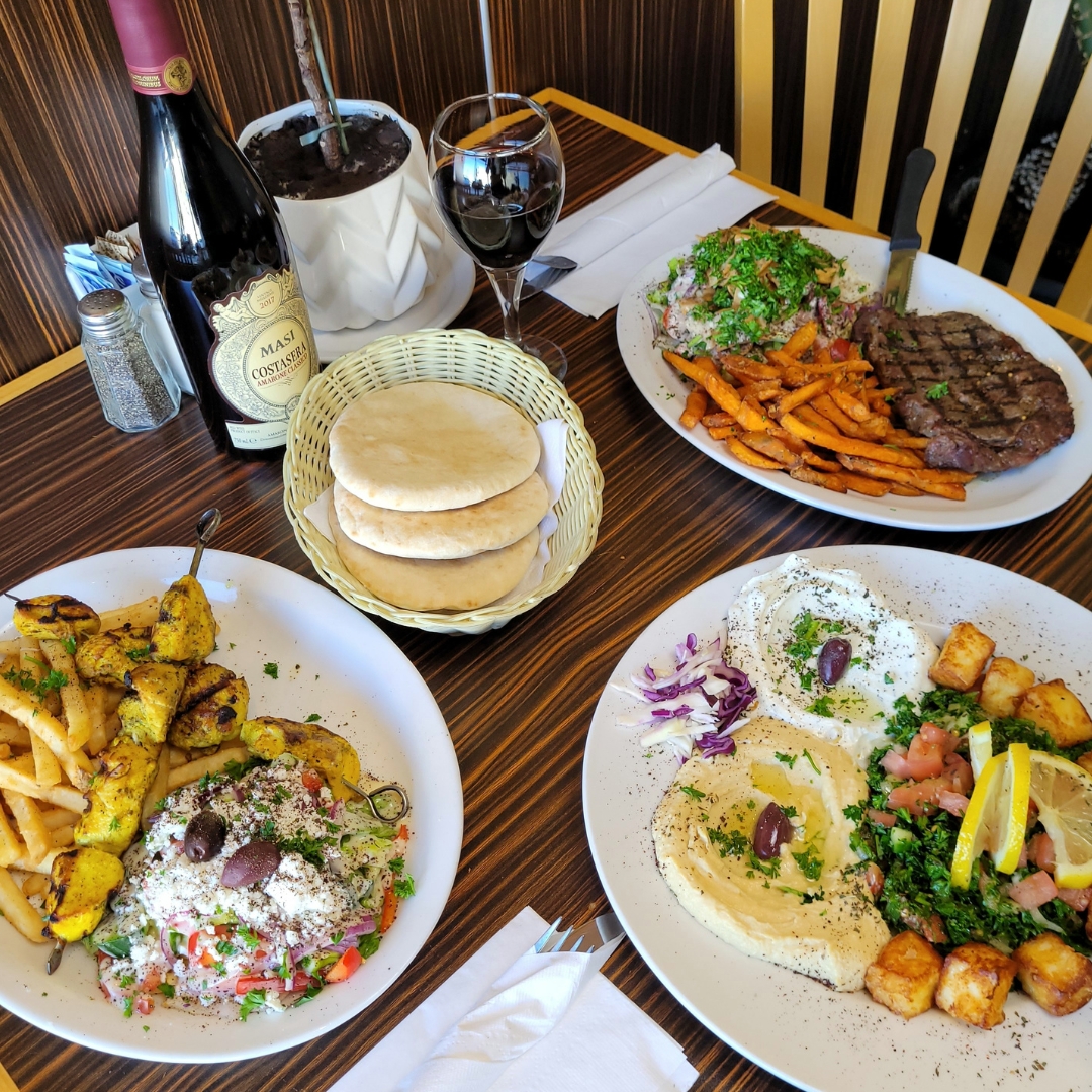 Table with Mediterranean dishes including grilled chicken skewers with fries, steak with sweet potato fries and salad, a plate with hummus, pita bread, and a glass and bottle of red wine.