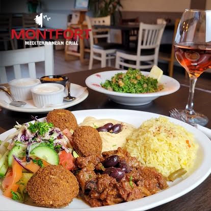 Plate of Mediterranean food with falafel, yellow rice, hummus, salad, and a glass of rosé wine in a restaurant setting with Montfort Mediterranean Grill logo.