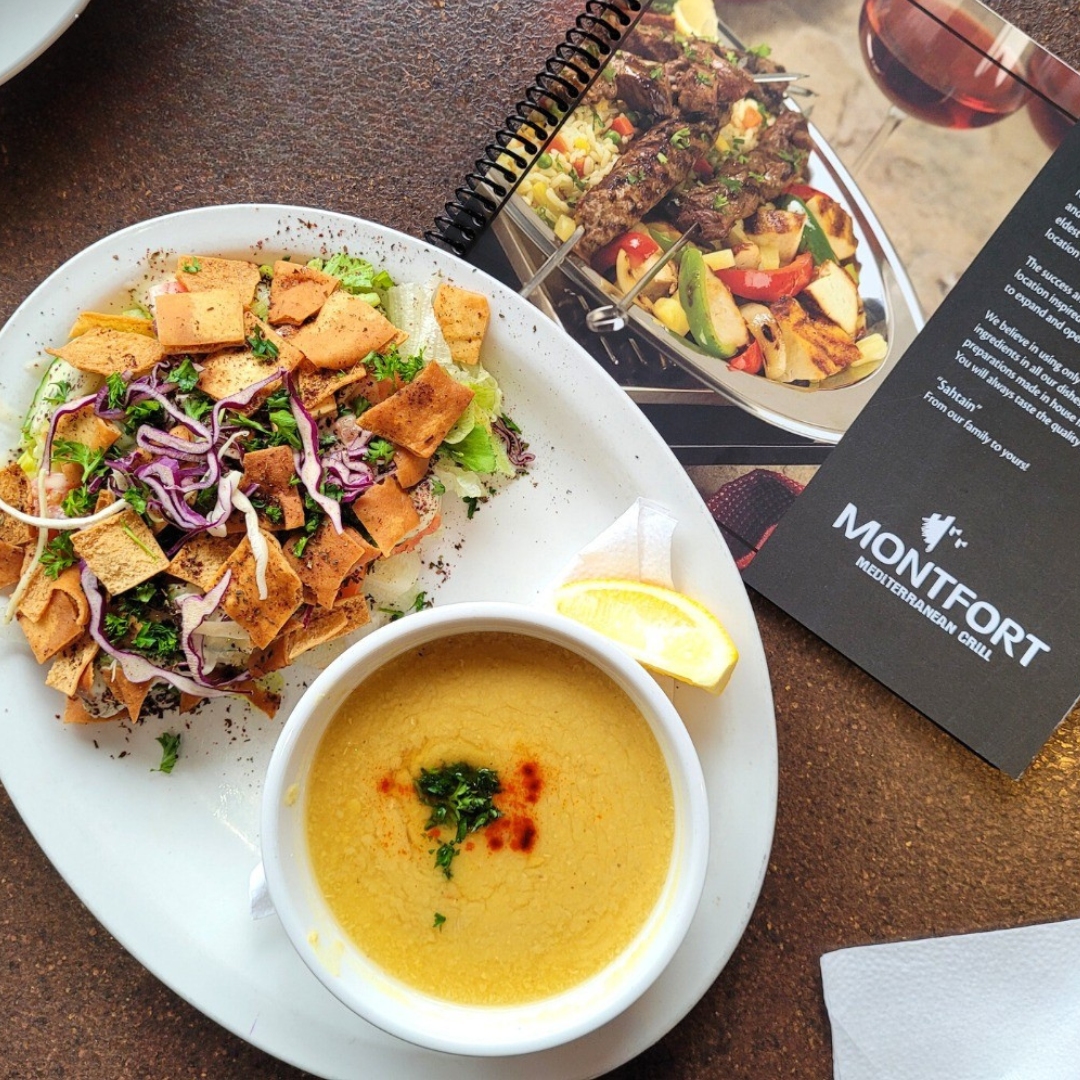 Plate with soup garnished with herbs and paprika, salad topped with toasted pita chips, and a lemon wedge, beside a Montfort Mediterranean Grill menu.
