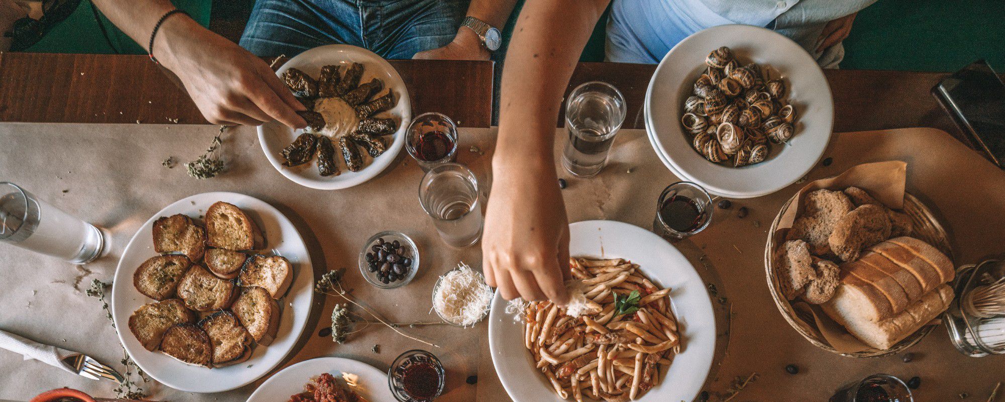 Table with plates of toasted bread, stuffed grape leaves with sauce, escargot, pasta with tomato sauce and cheese, a basket of bread, and glasses of water and red wine.