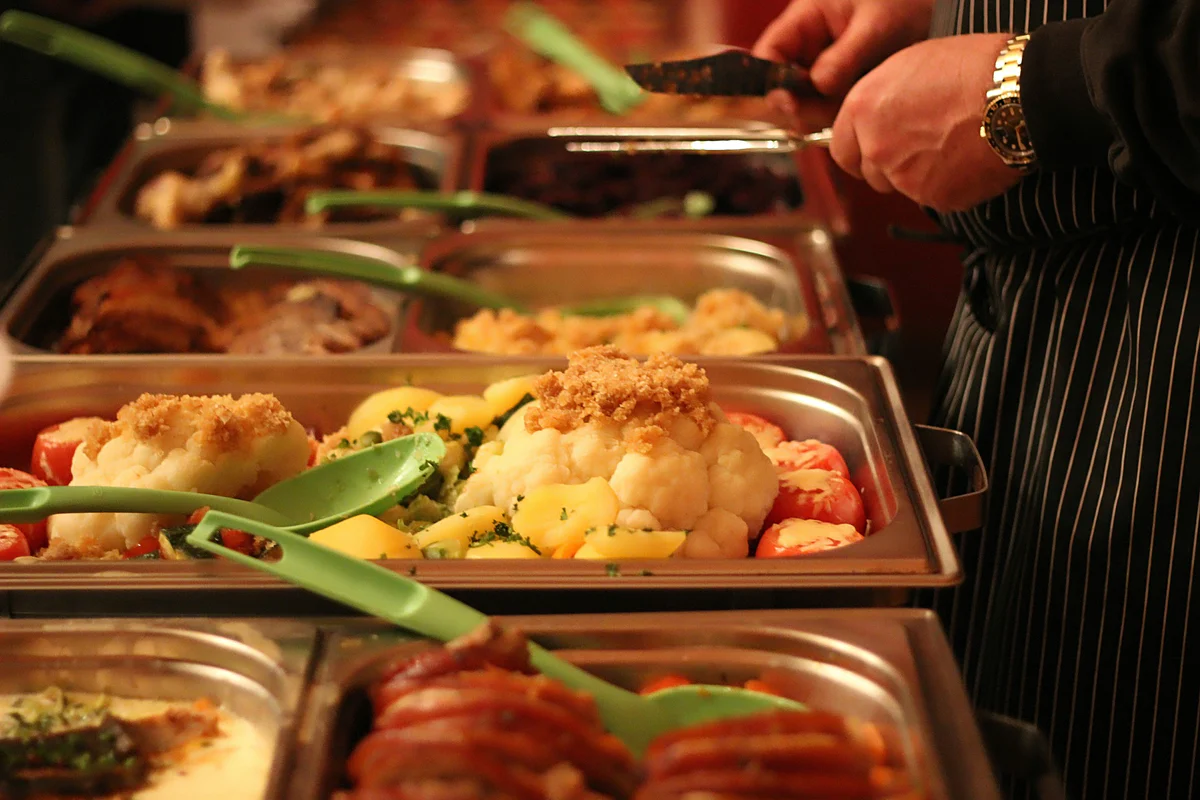 Buffet trays filled with various cooked dishes including cauliflower with crumbs, vegetables, and meat, with a person serving food.