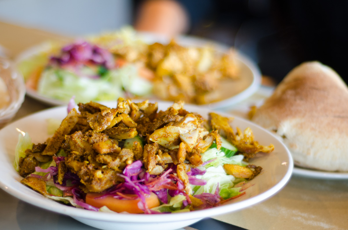 Plate of seasoned grilled chicken served over a salad with lettuce, purple cabbage, and tomato slices, with pita bread on the side.