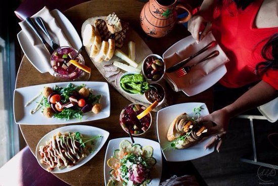 Overhead view of a wooden table with various tapas dishes including salad, bread, olives, and drinks, with a person in a red top reaching for food.