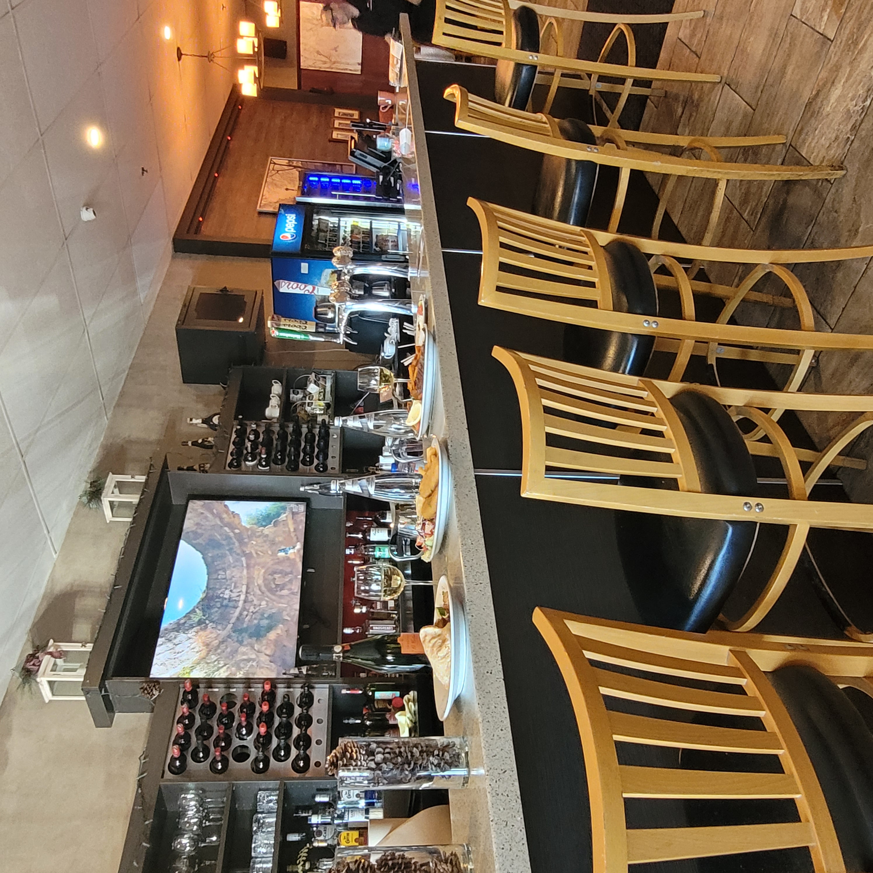 Bar counter with wooden stools, plates of food, wine glasses, wine bottles, and a TV mounted on the wall showing a stone structure.