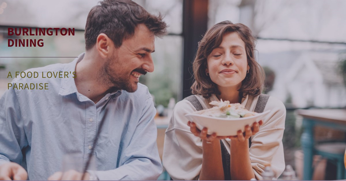 Smiling woman holding a plate of food close to her face with eyes closed, sitting beside a man in a light blue shirt at a dining table.