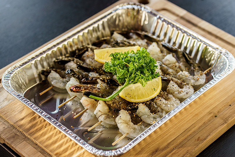 Tray of raw tiger shrimp skewers seasoned and garnished with lemon wedges and parsley on a wooden surface.