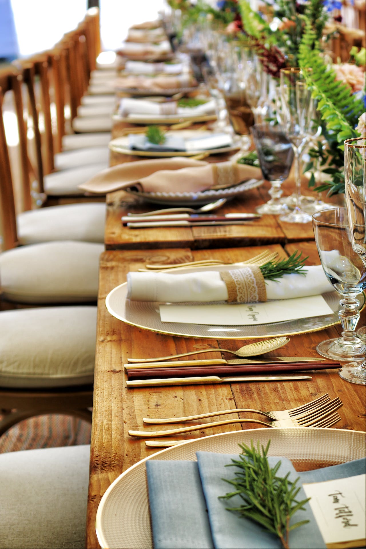 Long wooden dining table set with plates, cloth napkins wrapped with sprigs of rosemary, gold flatware, glassware, and greenery centerpieces.